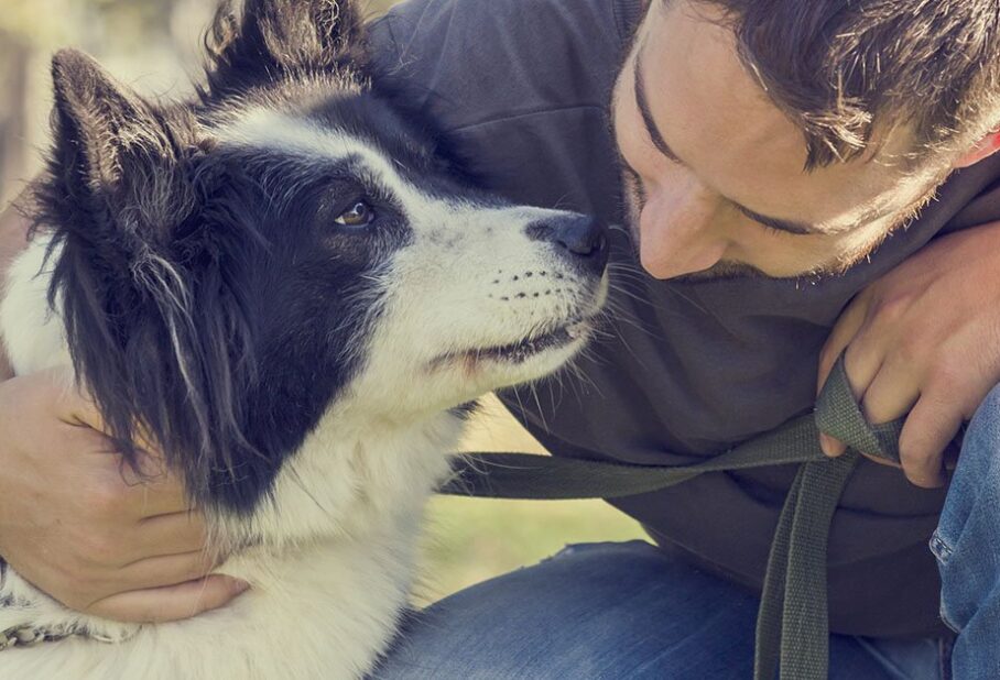 A person smiling and embracing a black and white dog in a park setting.
