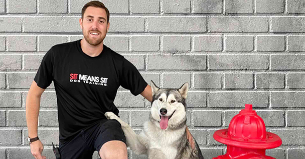 Man in a black shirt poses with a smiling husky beside a red fire hydrant against a gray brick wall.