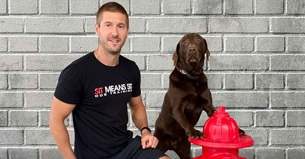 Man in a black shirt poses next to a brown dog beside a red fire hydrant, with a grey brick wall in the background.