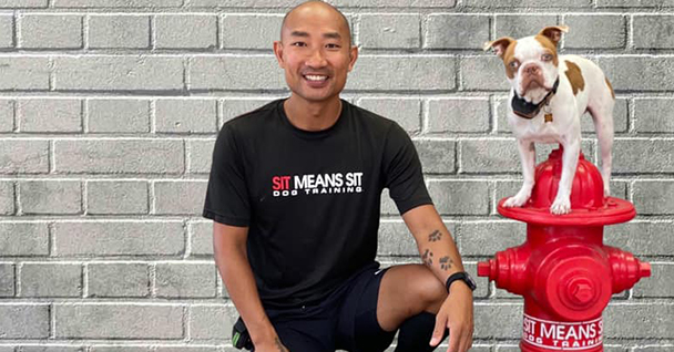 A person sits beside a dog perched on a red fire hydrant against a gray brick wall.
