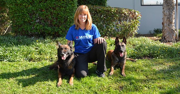 Woman kneeling on grass with two dogs, both looking happy and alert in a sunny outdoor setting.
