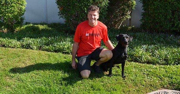 A man in a red shirt kneels beside a black dog on green grass.