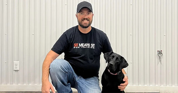 A man kneeling next to a black dog smiling for a picture