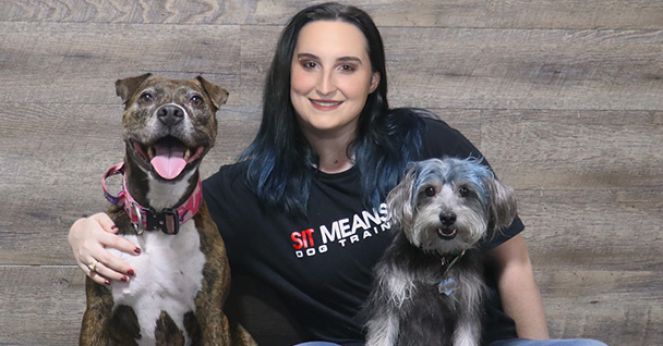 a woman sitting between with her arms around two dogs smiling for a photo