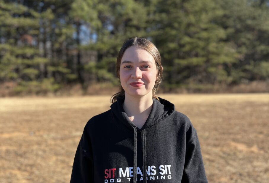 A person wearing a black hoodie stands outdoors with trees in the background on a clear day.