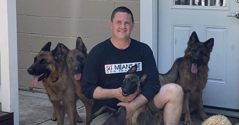 A man sits on the ground with four dogs, smiling and holding one of the dogs in his lap.