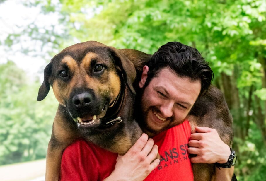 A man joyfully carries a large dog on his shoulders outdoors, surrounded by greenery.