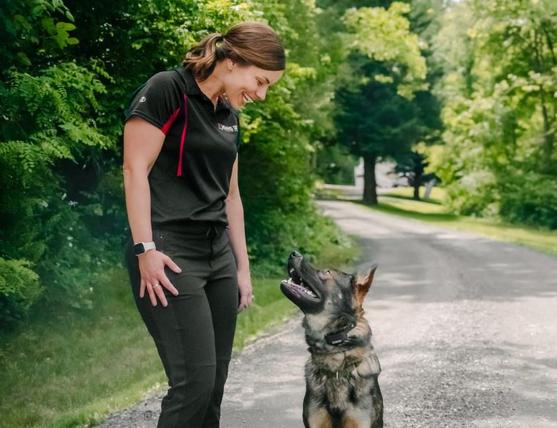 A woman smiles down at a German Shepherd on a wooded path.