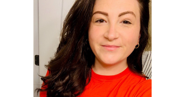 Woman with long brown hair smiling in a red shirt, standing in front of a bathroom mirror.