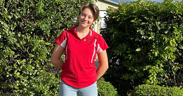 A smiling young person in a red shirt stands outdoors with green foliage in the background.