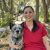A woman in a red shirt smiles next to her dog in a wooded area.