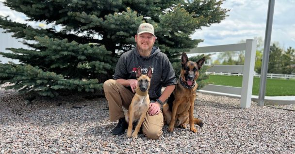 A man and two dogs sitting on the ground smiling for a picture with tree in the background