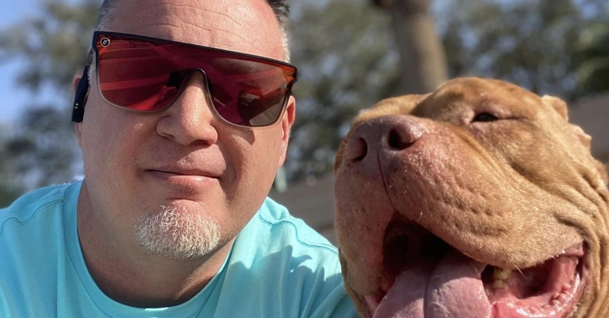 A man with sunglasses smiles next to a happy dog outdoors, with palm trees in the background.