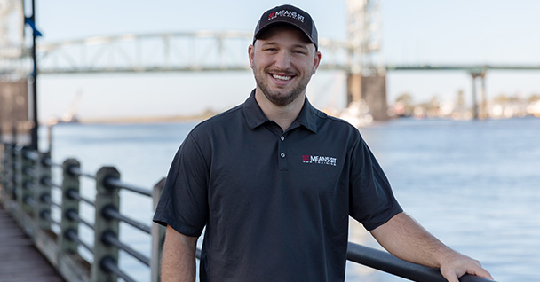 Smiling man in a branded shirt stands by a river with a bridge in the background.